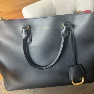 Svart handväska från Ralph Lauren - Snygg svart handväska i skinn från Ralph Lauren med guldfärgade metalldetaljer och rödfärgat foder. Väskan har dubbla handtag, innerfack med dragkedja och en stilren, klassisk design som passar till det mesta. Köpt för 3600 
