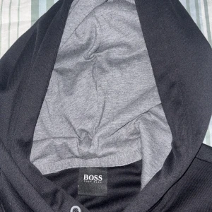 Svart hoodie från BOSS med logga - Snygg svart hoodie från BOSS med stor broderad logga framtill. Tröjan har huva med grå insida och svarta snören för justering. Perfekt för dig som gillar stilrena och exklusiva plagg med streetkänsla. helt ny ey anvönd pga storleken