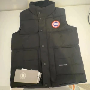 Svart dunväst från Canada Goose - Svart dunväst från Canada Goose med hög krage, två stora fickor framtill och klassisk logotyp-patch på bröstet. Västen har tryckknappar och är perfekt för lager-på-lager. Snygg och stilren design som funkar till det mesta.