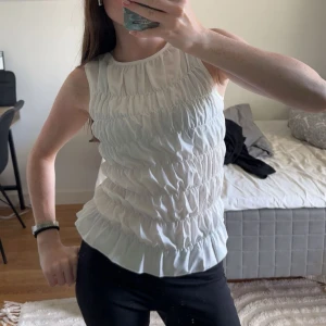 Vit ärmlös blus med volang - Supersöt vit blus med ärmlös design och volangkant nertill. Toppen har ett rynkat tyg i flera vågräta partier som ger en cool struktur och luftig känsla. Perfekt att styla med jeans eller kjol för en fräsch look.