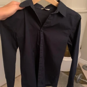 Mörkblå skjorta från C, stl 164 - Snygg mörkblå skjorta från C i storlek 164. Klassisk modell med krage, långa ärmar och knäppning framtill. Perfekt för dig som gillar en stilren look. Skjortan är tillverkad i ett mjukt och bekvämt bomullstyg.