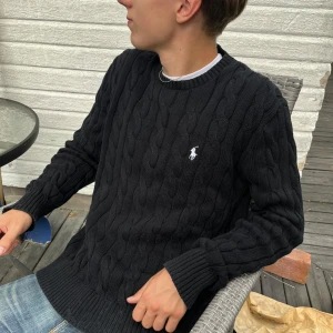 Svart kabelstickad tröja Polo Ralph Lauren - Kabelstickad svart tröja från Polo Ralph Lauren med klassisk rund halsringning och broderad vit logga på bröstet. Tröjan är långärmad och har ribbade muddar vid ärmslut och nederkant. Perfekt för dig som gillar stilren och tidlös design.