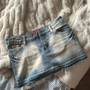 Blå jeanskjol med nitar från Reverb - Snygg kort jeanskjol från Reverb Jeans i ljusblå tvätt med slitna kanter. Baksidan har broderad rosa M och detaljer på bakfickorna. Klassisk femficksmodell med bälteshällor och dragkedja. Liten i storlek!