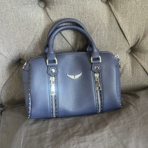 Blå handväska med silverdetaljer - Snygg blå handväska i skinn med två rejäla handtag och coola silverfärgade dragkedjor framtill. Väskan har en stilren form och ett litet silverfärgat emblem på framsidan. Perfekt för dig som vill ha en väska med edge och stil.