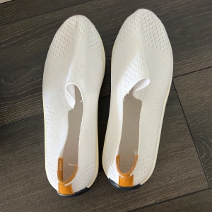 Vita stickade slip-on sneakers - Snygga vita slip-on sneakers med stickad ovandel och coola detaljer i brunt skinn baktill. Sulan är chunky med kontrastfärger i rött och mörkblått. Perfekt för dig som gillar minimalistisk och modern stil. Lätta och bekväma att dra på sig.