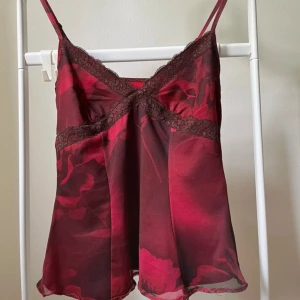 rött blommigt linne från Urban Outfitters - Supersött rött linne från Urban Outfitters med blommigt mönster och tunna axelband. Toppen har spetsdetaljer vid bysten och en lätt glansig finish. Perfekt att styla med jeans eller kjol för en trendig look.