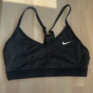 Svart glittrig sporttopp från Nike - Snygg svart sporttopp från Nike med glittriga detaljer och smala axelband. Toppen har en klassisk v-ringning och ett vitt Nike-märke framtill. Perfekt för träning och ger en sportig vibe med extra bling.