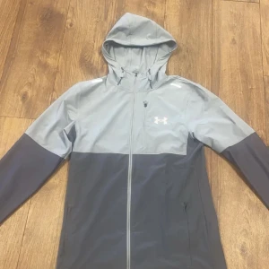 Under Armour grå vindjacka med huva - Snygg tvåfärgad vindjacka från Under Armour i ljusgrått och mörkgrått. Jackan har dragkedja vid bröstet och luva ny pris 1200
