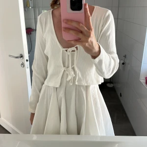 Vit ribbad kofta med snörning från Monki - Supersöt vit ribbad kofta från Monki i croppad modell med långa ärmar och snygg snörning framtill. Perfekt att slänga över en klänning eller linne för en chill och trendig look. Tunn och mjuk, passar grymt till vår och sommar.