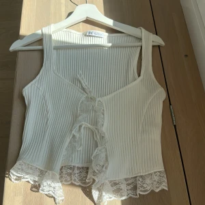 Vit ribbad linnetopp från Zara - Supersöt vit ribbad linnetopp från Zara med spetskant nertill och snörning framtill. Toppen är ärmlös och har breda axelband. Perfekt för dig som gillar romantiska detaljer och vill ha en feminin look.