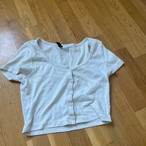 Vit croppad topp med knappar H&M Divided - Superfin vit croppad topp från H&M Divided med korta ärmar och knappar framtill. Toppen har en djup rundad urringning och är gjord i mjuk bomull. Perfekt till jeans eller kjol för en chill och enkel look.