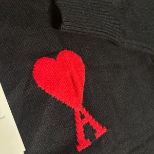 Svart stickad tröja från Ami Paris - Svart stickad tröja från Ami Paris med ikoniskt rött hjärta och A-logga på bröstet. Tröjan har rund halsringning, ribbade muddar och är långärmad. Perfekt för dig som vill ha en stilren men ändå unik look.