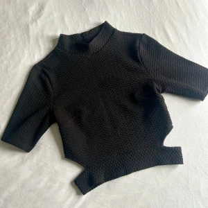 Svart strukturerad polotopp - Cool svart polotopp med unik struktur och asymmetrisk nederkant. Toppen har halvärmade ärmar och är tillverkad i ett stretchigt material med mönstrad yta som ger en modern vibe. Perfekt för dig som gillar edgy detaljer och vill sticka ut.