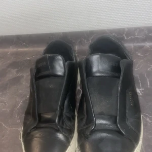 Svarta sneakers från Axel Arigato - Svarta sneakers från Axel Arigato i skinn med vit platt sula. Diskret logga i guld på sidan och stilren design utan snörning. Perfekta för dig som gillar minimalistisk och clean stil.