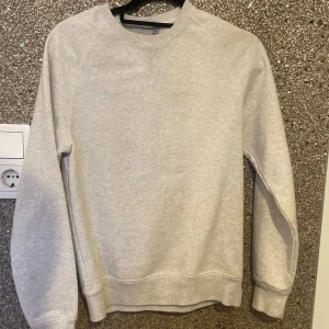 sweatshirt från Carhartt  - Säljer en stilren ljusgrå sweatshirt från Carhartt i storlek XS. Tröjan har rund hals, ribbade muddar och en liten gul Carhartt-logga broderad på ärmen. Fint skick!