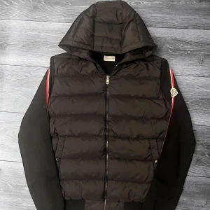 Svart dunväst med huva från Moncler - Svart dunväst från Moncler med huva och dragkedja framtill. Västen har quiltad design, svarta ärmar med röda och vita ränder längs axlarna samt Moncler-logga på vänster ärm. Perfekt för lager-på-lager och streetstyle.