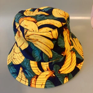 🍒Bucket hat med bananmönster🍒 - 🍒BANANA🍒Färgglad bucket hat med coolt bananmönster i gult och grönt mot mörkblå botten. Hatten har brett brätte och är tillverkad i mjukt tyg, perfekt för att sticka ut i mängden. Snygg accessoar för dig som gillar lekfull stil och tropiska vibes! 🍒