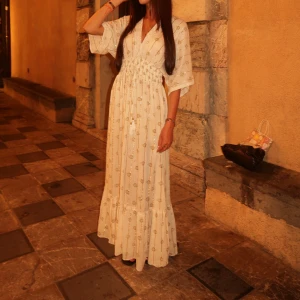 Ljus långklänning med guldmönster - Superfin ljus långklänning i bohemisk stil med guldiga detaljer och diskret mönster. Klänningen har v-ringning, vida trekvartsärmar och markerad midja med smock. Perfekt för sommarkvällar och ger en romantisk vibe.