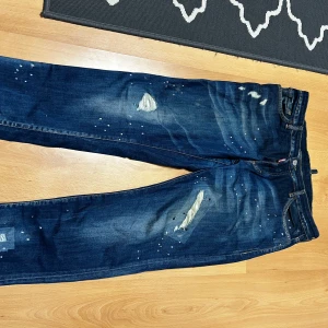 Dsquared2 blå distressed jeans - Säljer ett par blå jeans från Dsquared2 i storlek 52. Jeansen har en cool distressed look med slitningar, färgstänk och lagade detaljer. Klassisk femficksmodell med raka ben och snygg tvätt. Perfekta för dig som gillar streetstyle och vill sticka ut.
