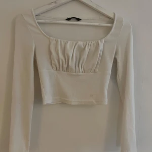 Vit croppad långärmad topp - Snygg vit croppad topp med långa ärmar och fyrkantig ringning. Framsidan har en rynkad detalj över bysten som ger en cool och trendig look. Tillverkad i ett mjukt, ribbat material som sitter skönt på kroppen.