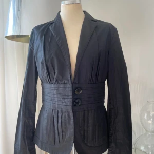 Svart kavaj med markerad midja - Snygg svart kavaj med bred, veckad midjedetalj och två stora knappar framtill. Jackan har långa ärmar, klassisk krage och plisserade detaljer både fram och bak. Perfekt för dig som vill ha en stilren men unik look.