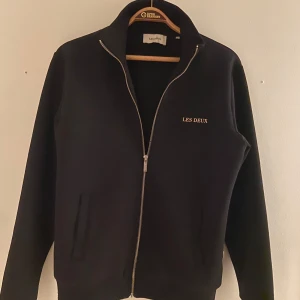 Svart zipjacka från Les Deux - Svart jacka från Les Deux med stilren design och guldfärgad logga på bröstet. Jackan har hel dragkedja, ståkrage och två fickor framtill. Tillverkad i mjukt material och passar perfekt till en clean och avslappnad look.
