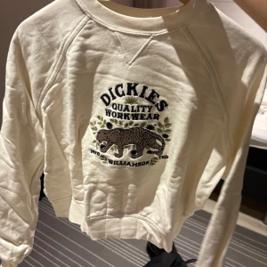 Vit Dickies sweatshirt med leopardbrodyr - Säljer en vit sweatshirt från Dickies i storlek XS. Tröjan har broderad text och en leopard på bröstet, samt coola detaljer med blad runt motivet. Mjuk bomullskänsla och långa ärmar, perfekt för en chill och stilren look.
