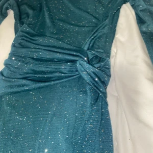 Mörkblå glittrig långklänning - Snygg mörkgrön långklänning med glittriga detaljer och lång ärm. Klänningen har en twistad detalj i midjan och en slits nertill för extra vibe. Perfekt för dig som vill sticka ut med lite extra glitter och glam.