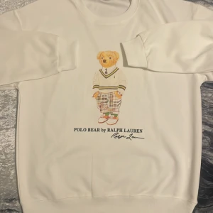 Vit Polo Bear sweatshirt från Ralph Lauren - Vit sweatshirt från Polo Ralph Lauren med ikoniskt Polo Bear-tryck framtill. Tröjan har rund hals, långa ärmar och ribbade muddar. Perfekt för dig som gillar klassisk och lekfull stil. Mjuk och skön bomullskänsla.