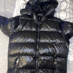 Svart pufferjacka från Moncler L - Snygg svart pufferjacka från Moncler i storlek L. Jackan har glansig finish, huva, två fickor med dragkedja och klassisk Moncler-logga på bröstet. Perfekt för kalla dagar när du vill hålla stilen och värmen.