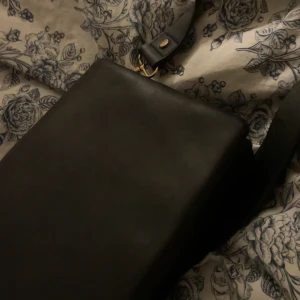 Svart axelväska i skinn med gulddetaljer - Stilren svart axelväska i slätt skinn med bred axelrem och snygga guldfärgade metalldetaljer. Väskan har magnetlås och dragkedja på insidan för extra säkerhet. Perfekt för dig som gillar minimalistisk och klassisk design.