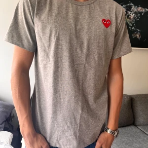 Grå Comme des Garçons Play t-shirt - Grå t-shirt från Comme des Garçons Play med det ikoniska röda hjärtat med ögon broderat på bröstet. Klassisk passform, rund hals och korta ärmar. Perfekt basic med en twist för dig som gillar streetstyle.