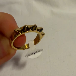 Guldfärgad ring med nitar, storlek 7 - Cool guldfärgad ring i storlek 7 med fem spetsiga nitar runt ovansidan. Ringen har en chunky känsla och ger en edgy vibe till din stil. Perfekt för dig som gillar statement-smycken och vill sticka ut lite extra.