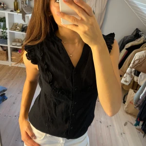 Svart broderad volangtopp  - Supersöt svart blus från Cache Cache med volangdetaljer längs axlar och bröst. Ärmlös modell med knappar framtill och lätt transparent tyg. Perfekt att styla med jeans eller kjol för en trendig look.