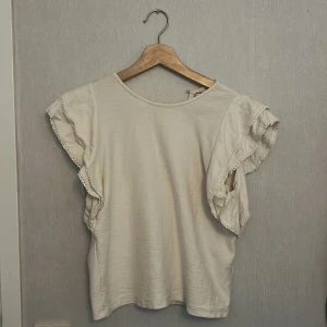 Vit volangtopp Holly & Whyte XS - Supersöt vit topp från Holly & Whyte med volangprydda ärmar och fina spetsdetaljer. Toppen har rund halsringning fram och v-ringning bak, vilket ger en unik look. Perfekt för dig som gillar romantiska och luftiga plagg.