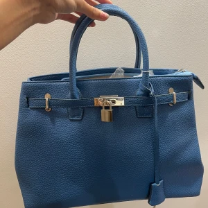 Blå handväska i skinn med låsdetalj (birkin) - Snygg blå handväska i strukturerat skinn med två handtag och cool låsdetalj i silver framtill. Väskan har metallfötter undertill och klassisk boxig form. Perfekt för dig som vill ha en stilren och trendig accessoar med lyxig känsla. Köpt second hand så osäker på om det är en äkta, därav priset. 