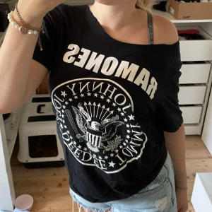 Svart Ramones t-shirt offshoulder - Svart t-shirt med ikoniskt Ramones-tryck i vitt framtill. Modellen är offshoulder och har korta ärmar, vilket ger en avslappnad och cool vibe. Tillverkad i mjuk bomull, perfekt för dig som gillar bandtröjor och streetstyle. Pris går att diskuteras.