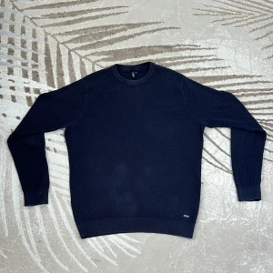 Mörkblå stickad tröja Hampton Republic - Mörkblå stickad tröja från Hampton Republic 27 med diskret rutstruktur i materialet. Klassisk rund halsringning och ribbade muddar vid ärmslut och nederkant. Perfekt för dig som gillar stilren och enkel design.
