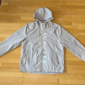 Moncler Windbreaker - Snygg ljusgrå vindjacka från Moncler i storlek XL. Jackan har huva, dragkedja framtill, tre fickor med dragkedja och stort Moncler-tryck på bröstet. Tillverkad i lätt och smidigt syntetmaterial, perfekt för blåsiga dagar. Storleken sitter nästan som L. Ingen prislapp. Skickas så fort som möjligt!⚡️