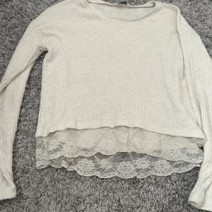 Vit ribbad tröja med spetsdetalj - Supersöt vit ribbad tröja med lång ärm och bred spetskant nertill. Tröjan har en rund halsringning och den genomskinliga spetsen med blommönster ger en extra touch. Perfekt för dig som gillar romantiska detaljer och en chill stil.