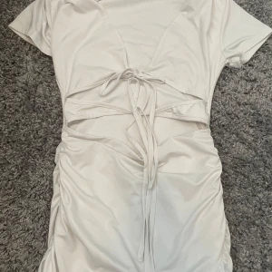 Vit kort klänning med snörning - Snygg vit kortklänning med trendig snörning framtill och cut out-detaljer i midjan. Klänningen har korta ärmar och en figurnära passform som framhäver siluetten. Perfekt för dig som vill sticka ut med en modern look.