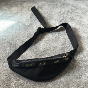 Svart magväska från BOSS Hugo Boss - Snygg svart magväska från BOSS Hugo Boss med justerbart band och guldfärgade dragkedjor. Väskan har logga på framsidan och flera fack med dragkedja, perfekt för att bära det viktigaste. Tillverkad i slitstark polyester med stilren design.