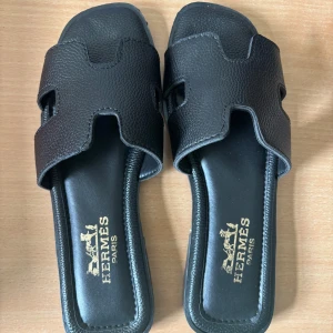 Svarta sandaler - Svarta sandaler med ikonisk H-design på ovandelen. Tillverkade i präglat skinn med platt sula och öppen tå. Snygg minimalistisk look, perfekt för varma dagar. Levereras i klassisk orange box