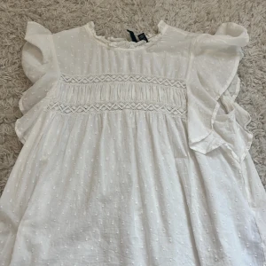 Vit somrig blus från Vero Moda - Supersöt vit blus från Vero Moda i bomull med broderade prickar och spetsdetaljer. Volangärmar och romantisk känsla, perfekt för varma dagar. Lätt och luftig modell som ger en fräsch vibe.