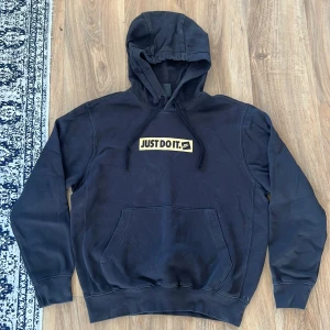 Svart Nike hoodie med Just Do It-tryck - Svart hoodie från Nike med klassiskt guldigt 'Just Do It.'-tryck på bröstet. Tröjan har huva med snörning och en stor känguruficka framtill. Såklart tvättad innan den skickas