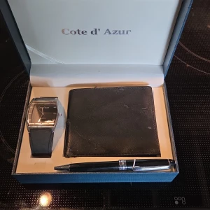 Svart klocka från Cote d'Azur - Stilren svart klocka från Cote d'Azur med rektangulärt silverfärgat boett och svart urtavla. Klockan har ett svart armband och minimalistisk design, perfekt för dig som gillar en clean look. Levereras i presentbox tillsammans med matchande plånbok och penna.Plånboken blivit lite skavde aldrig använt klocka skyddsplast kvar     