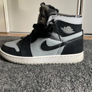 Nike Air Jordan 1 Zoom Air svart/grå - Nike Air Jordan 1 Zoom Air i svart och grått med högt skaft och coola detaljer. Ovandel i mocka och skinn, klassisk Swoosh-logga på sidan och Air Jordan Wings på ankeln. Tungan har ett mönster i svart och grått samt Zoom Air-logga. Perfekta för dig som gillar streetstyle. Det slitna på toppen är designen