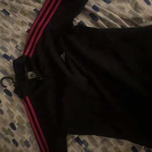 Svart Adidas jacka med rosa ränder - Svart Adidas jacka med klassiska rosa ränder längs ärmarna och liten logga på bröstet. Jackan har lång ärm, dragkedja framtill och sportig vibe. Perfekt för dig som gillar streetstyle och vill sticka ut med färgdetaljer.