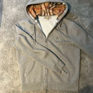 Grå Burberry hoodie med dragkedja - Snygg grå hoodie från Burberry med klassiskt rutigt foder i huvan. Tröjan har dragkedja framtill, snörning i huvan och två fickor. Tillverkad i mjuk bomull och polyesterblandning. Perfekt för dig som vill ha en stilren och lyxig vardagslook.