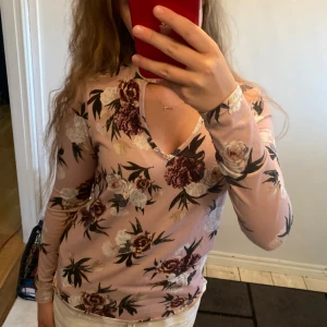 Blommig rosa långärmad topp - Supersöt långärmad topp i ljusrosa med blommigt mönster i vitt, vinrött och grönt. V-ringad med en liten cutout vid halsen som ger en extra detalj. Mjuk och stretchig, perfekt till jeans eller kjol.